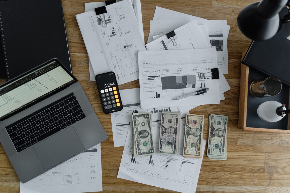 Money, finance documents, digital calculator, and a laptop spread out on a wooden desk.