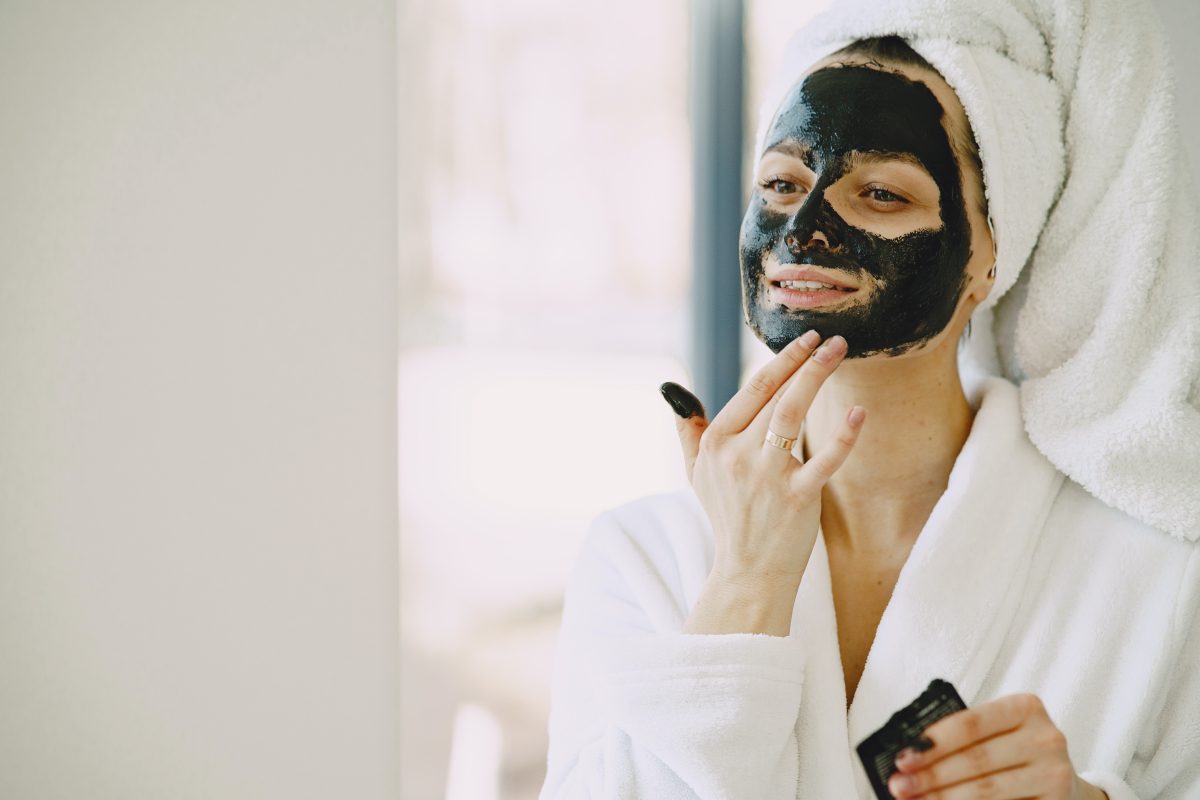 A woman wearing a white bathrobe and towel applying a black clay mask on her face