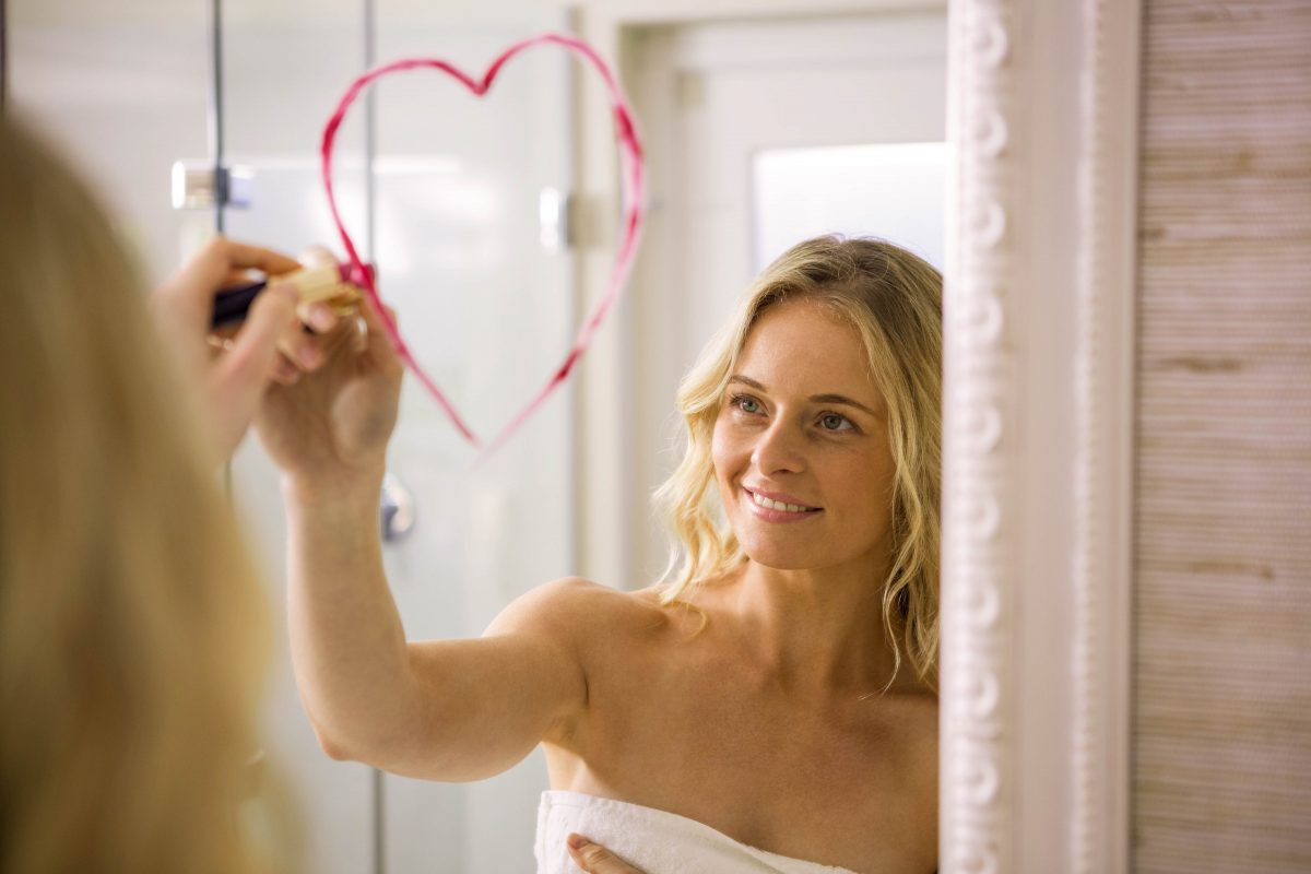 Blonde woman in a white towel drawing a heart on a mirror with red lipstick.