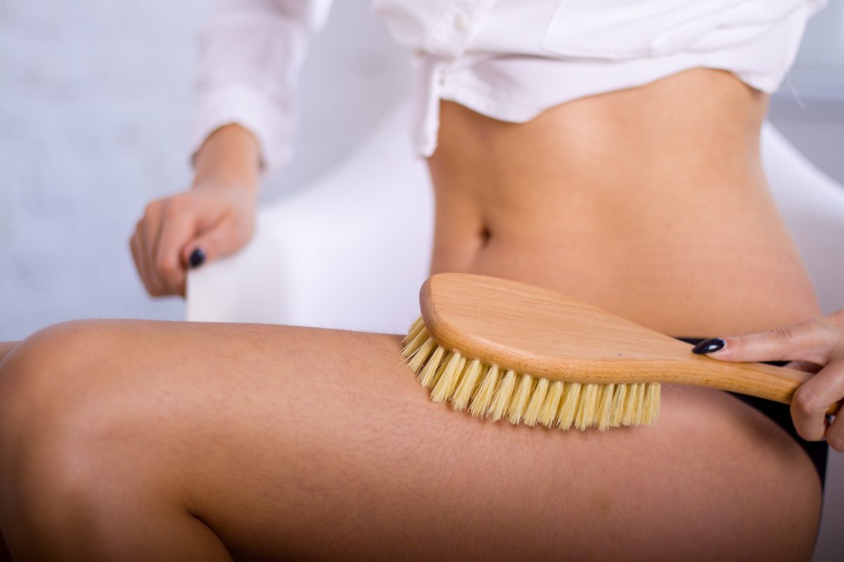 Close-up shot of a woman in black underwear dry brushing her upper thigh with a wooden brush, as part of her glow up routine.
