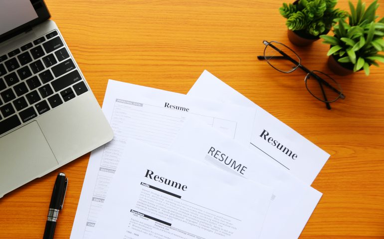 Several resumes on a wooden work desk with a pair of glasses and a laptop.