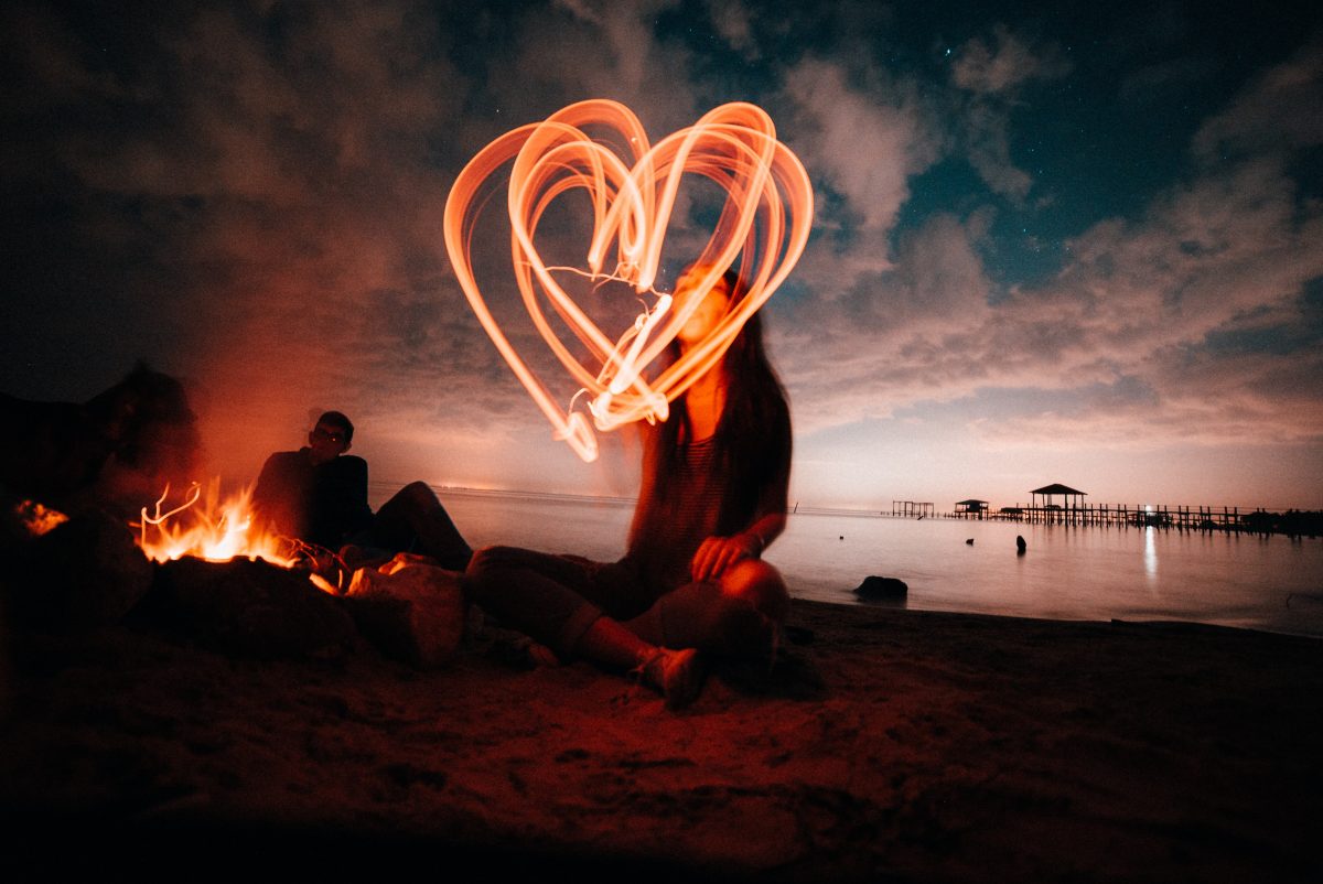 Creative shot of a long exposure heart with a woman sitting on a beach near a bonfire. 
