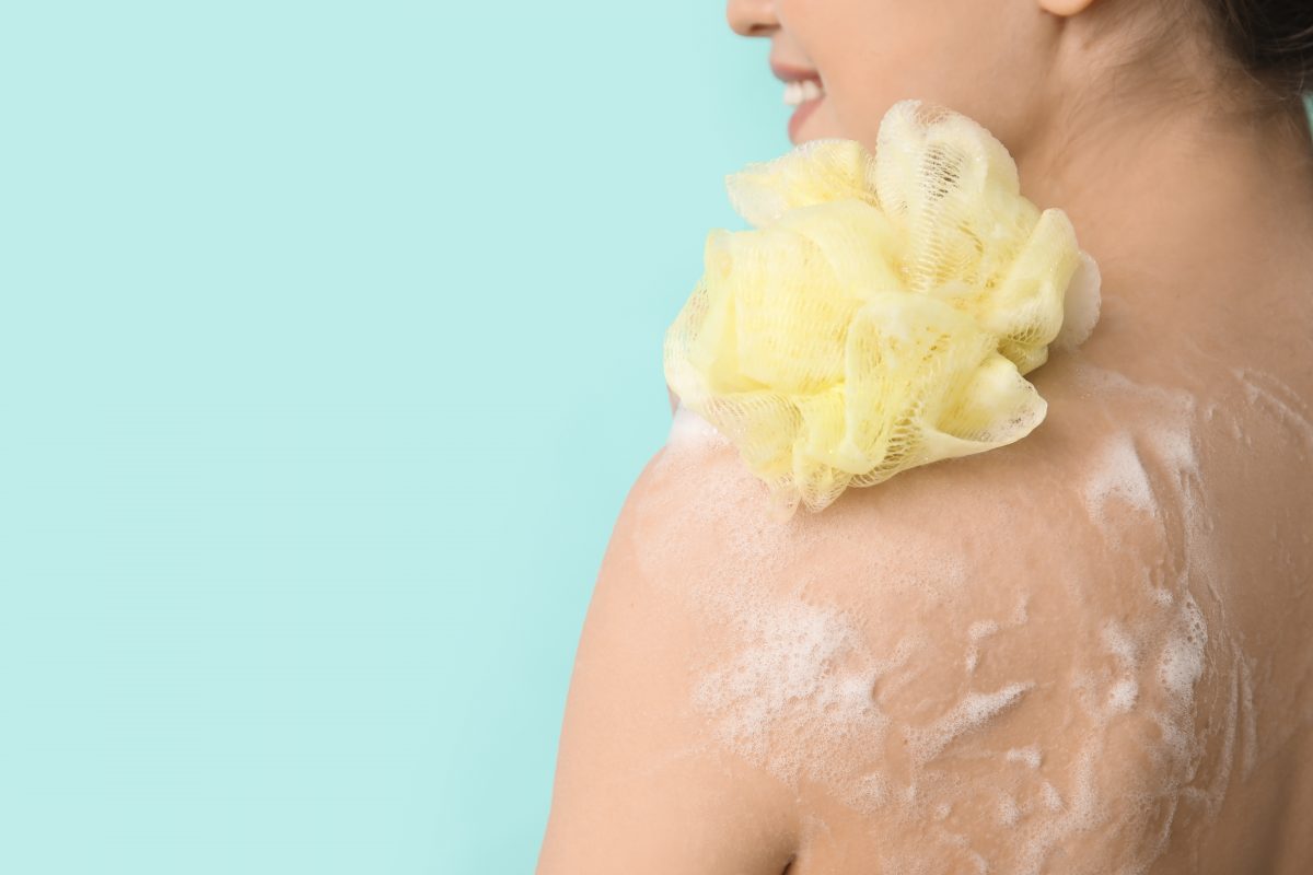 Close-up shot of a woman taking shower, holding a luffa sponge to her sudsy shoulder, part of her glow up routine.