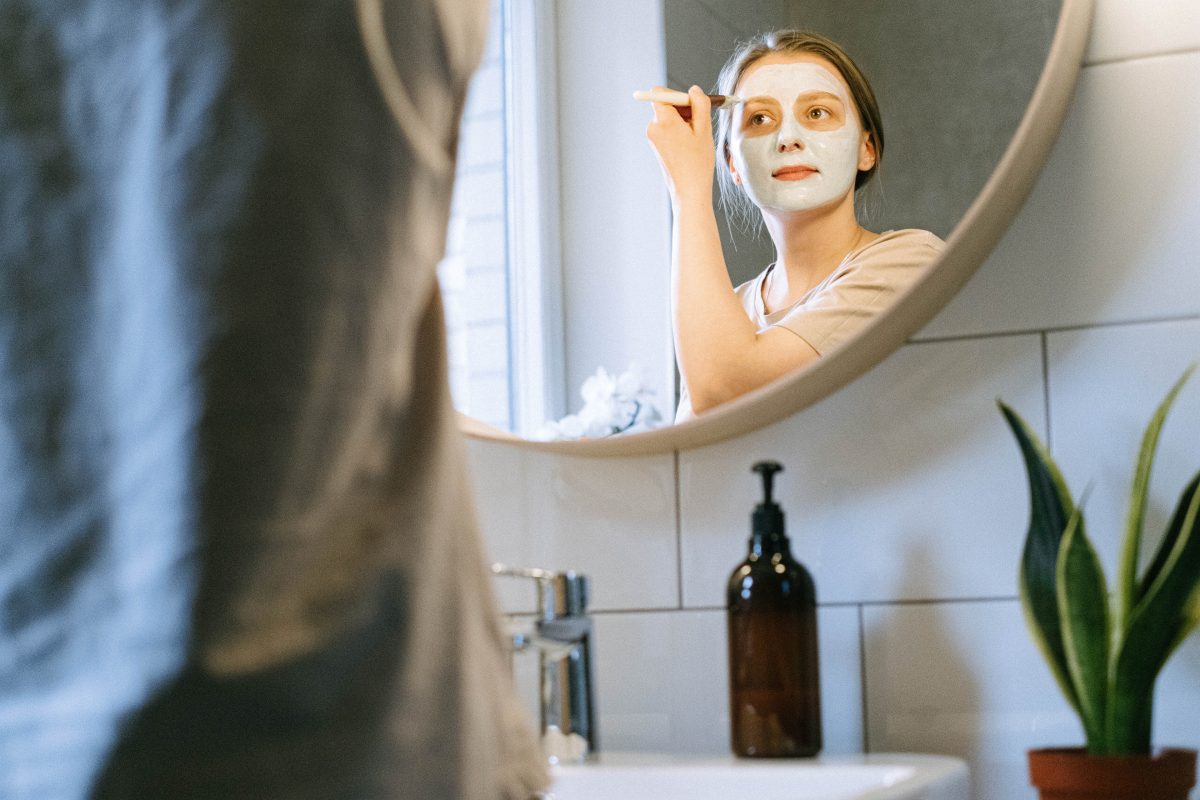 Perspective shot of a woman in a bathroom mirror applying a face mask, one of the ways on how to glow up instantly.