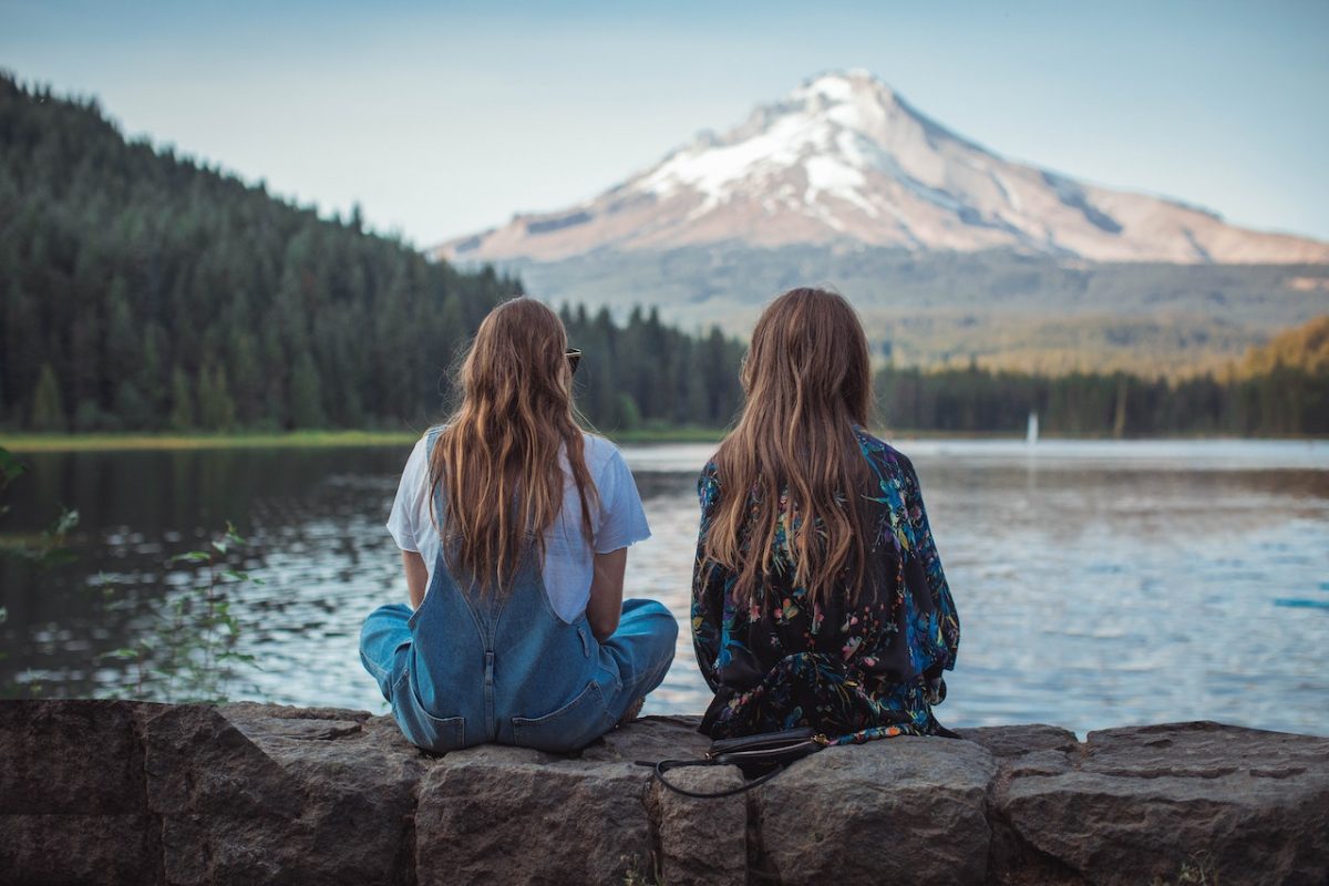 Two Women Sitting on Rock Facing on Body of Water and Mountain.